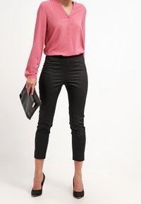 Femme portant un chemisier rose à manches longues, un pantalon noir court, des talons noirs, tenant une pochette noire sur fond blanc.