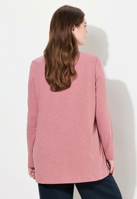 Maglietta a maniche lunghe in morbido tessuto rosa, con una vestibilità rilassata, orlo curvo e texture minima, abbinata a pantaloni scuri, vista da dietro.
