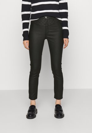 Vrouw draagt zwarte doorgestikte broek met hoge taille, zwarte loafers en een zwart-wit gestreepte trui met lange mouwen, staand op een grijze vloer.