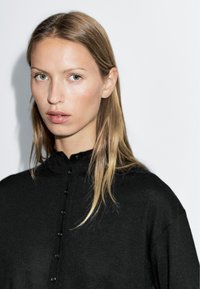 Black button-up shirt made of soft fabric, featuring a high collar and a relaxed fit. Model has straight blonde hair and light green eyes.