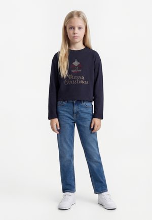 Ung jente står, iført svart genser med lange ermer med juledekorasjon og teksten "God jul", blå jeans og hvite joggesko.