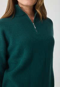 Σκούρο μπλε ζακέτα με ραβδώσεις, υψηλό λαιμό και κλείσιμο με φερμουάρ έως το ένα τέταρτο. Το ύφασμα έχει μαλακή υφή και άνετη εφαρμογή.