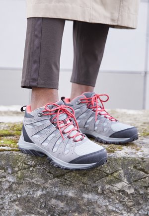 Persoon die grijze en roze wandelschoenen draagt, staand op een met mos bedekt stenen oppervlak met een bruine broek en een beige jas.