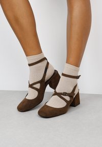 Zapatos de ante marrón con tacón bloque y detalles recortados, acompañados de calcetas ajustables, combinados con calcetines de canalé color crema.