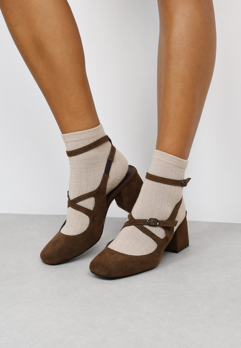 Zapatos de ante marrón con tacón bloque y detalles recortados, acompañados de calcetas ajustables, combinados con calcetines de canalé color crema.