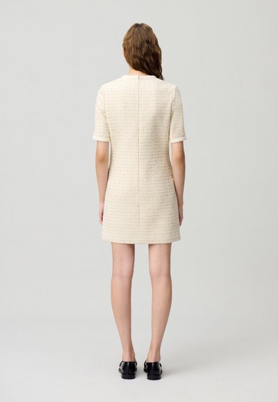 Robe courte en crème, texturée, avec des manches courtes et une fermeture éclair au dos. Portée avec des chaussures noires, mettant en valeur une silhouette ajustée.