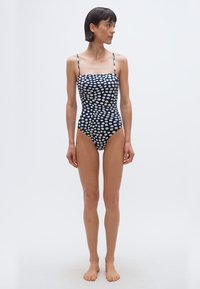 Jalakene paljajalu, kandes meresinist ühes tükis ujumistrikoodi valgete ebaregulaarselt jaotunud täppidega, vaadates vasakule tühja tausta taustal.