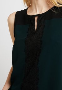 Top bez rękawów w ciemnozielonym kolorze, z czarnymi detalami i teksturowaną czarną koronkową wstawką z przodu. Zawiera element w kształcie dziurki na dekolcie.