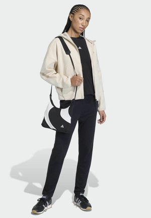 Crèmekleurig capuchonjack, zwart T-shirt, zwarte broek en zwarte sneakers. Het model draagt een schoudertas met een zwart-wit patroon.