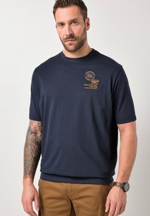 Mann trägt ein marineblaues kurzärmeliges T-Shirt mit einem luftfahrtbezogenen Logo auf der Brust, braune Hose, Armbanduhr und einen tätowierten linken Arm, steht vor weißem Hintergrund.