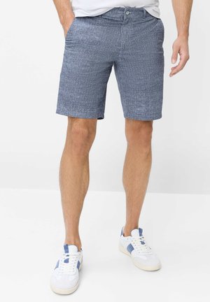 Mann trägt blau-weiß gestreifte Shorts, weiße Sneaker mit blauen Akzenten und ein weißes Hemd, die Hände in den Taschen.