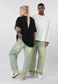 Γυναίκα με μακριά ξανθή αλογοουρά, φορώντας μαύρο πουκάμισο και ξεθωριασμένο τζιν, στέκεται σε πλάγια στάση· άνδρας με λευκό μακρυμάνικο πουκάμισο και ξεθωριασμένο τζιν κοιτάζει μπροστά.