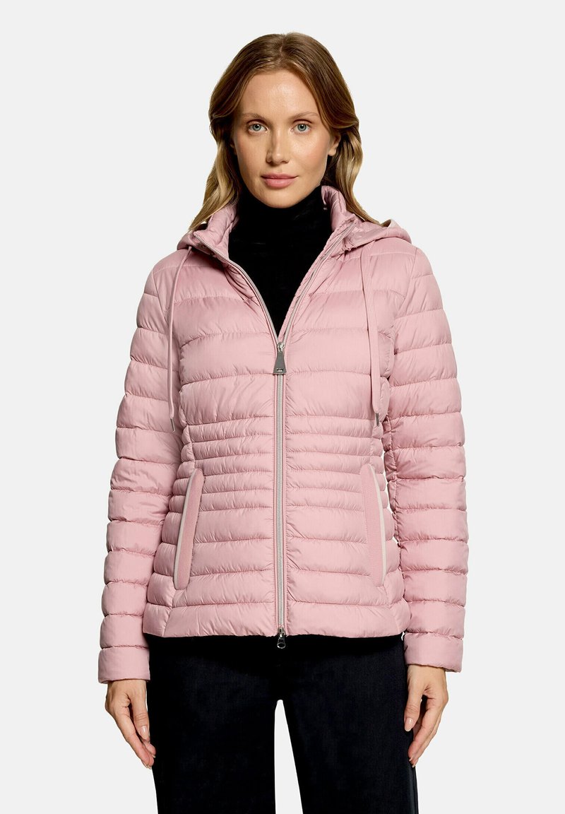 Femme portant une veste matelassée rose clair avec capuche et fermeture éclair devant un haut noir, debout devant un fond uni clair.