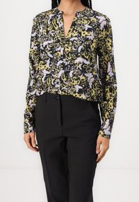 Femme portant un chemisier à manches longues noir, jaune et rose clair avec des fleurs, rentré dans un pantalon noir taille haute adapté.