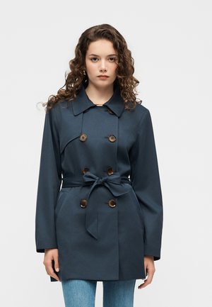 Mujer con cabello rizado lleva un abrigo trench azul oscuro de doble botonadura con cinturón, botones marrones y vaqueros azules sobre un fondo liso.