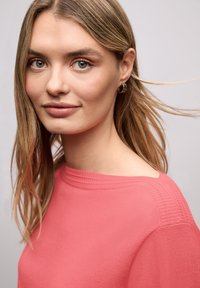 Młoda kobieta o jasnoblond włosach, nosząca złote kolczyki koła i koralowy sweter, patrząca łagodnie w obiektyw na neutralnym tle.