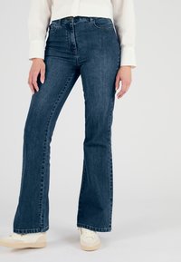 Vaqueros acampanados de color azul oscuro, elaborados en denim, que cuentan con una cintura alta, diseño de cinco bolsillos y sutiles detalles de costura a lo largo de las perneras.