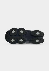 Σόλα σκοτεινού χρώματος παπουτσιού New Balance με μαύρο πέλμα και λευκές λαβές σε σχήμα διαμαντιού σε απλό γκρι φόντο.