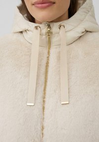 Lichtbeige nepbontjack met een gewatteerde capuchon, gouden rits en beige trekkoorden. Heeft een zachte textuur en een subtiele glans.