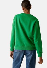 Felpa verde con scollatura a girocollo e polsini a coste, realizzata in tessuto morbido. Abbinata a jeans blu dal taglio rilassato, per un look informale.