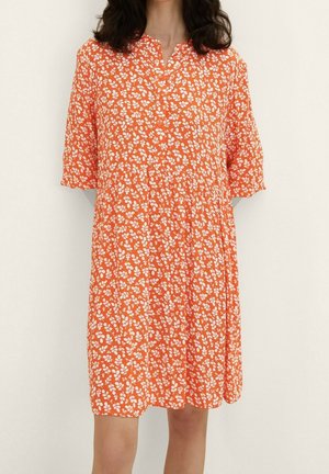 Nainen, joka käyttää polvipituista oranssia mekkoa, jossa on valkoinen kukkakuosi ja lyhyet hihat, seisoo vaaleaa yksinkertaista taustaa vasten.