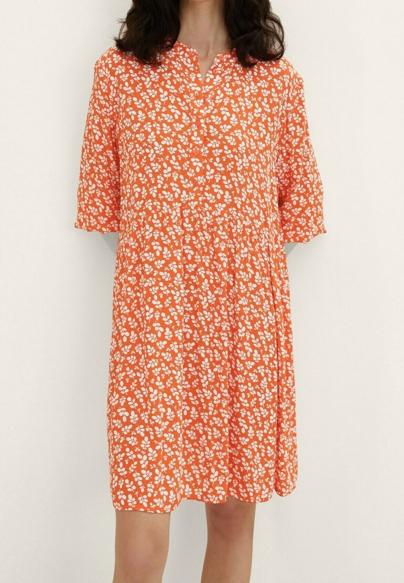 Femme portant une robe orange mi-longue à motif floral blanc et manches courtes, debout devant un fond clair uni.