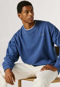 Νέος άνδρας καθισμένος σε ξύλινη καρέκλα, φορώντας μπλε φούτερ και λευκό παντελόνι, κοιτάζοντας προς τα δεξιά του με φόντο ένα απλό σκηνικό.