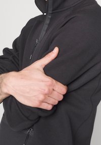 Chaqueta de forro polar negra con cremallera, que presenta un cuello alto, textura suave y detalles de costura, con las manos entrelazadas alrededor de la manga.
