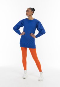 Kék, hosszú ujjú ruha puffos ujjakkal, narancssárga leggings-szel és fehér sportcipővel párosítva. Simaszerkezetű anyag, egyszerű dizájn és élénk színek.