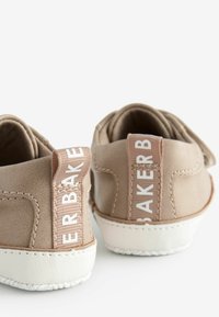 Tan suede kengät, joissa on valkoinen kumipohja. Ominaisuuksia ovat teksturoitu pinta ja takana oleva kangastagi, jossa lukee "BAKER." Minimalistinen muotoilu.