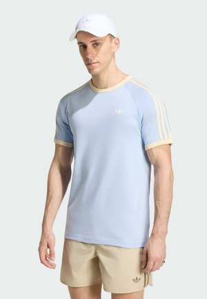 Mand iført lyseblå Adidas t-shirt med beige kant, beige Adidas shorts og hvid kasket, stående mod en ensfarvet lys baggrund.