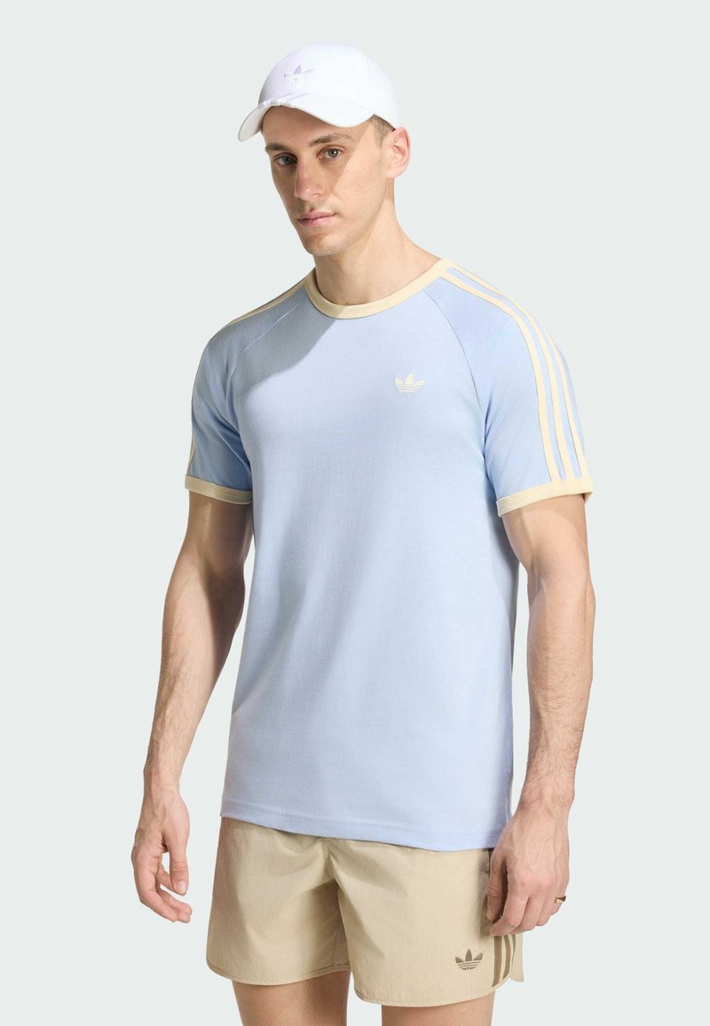 Uomo che indossa una t-shirt Adidas azzurro chiaro con bordatura beige, pantaloncini Adidas beige e cappellino bianco, in piedi davanti a uno sfondo chiaro uniforme.