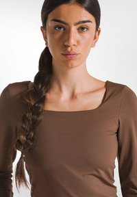 Maglietta marrone a maniche lunghe con scollatura quadrata, caratterizzata da una texture liscia e una vestibilità slim, abbinata a un dettaglio di capelli intrecciati sul lato sinistro.