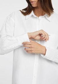 Camicia bianca con bottoni realizzata in tessuto morbido, con collo a punta, polsini regolabili e un piccolo logo a patch sulla tasca del petto.