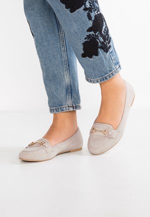 Fødder med lysegrå ruskindsløbesko med guldfarvet hestebid-detalje, kombineret med blå jeans med sort blomsterbroderi nær manchetterne.