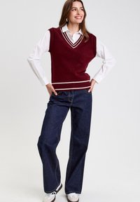 Gilet en tricot bordeaux avec un décolleté en V et des accents à rayures blanches, porté sur une chemise blanche à col, associé à un jean large bleu marine et des baskets blanches.
