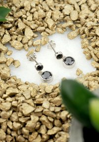 Silvriga hängande örhängen med runda svarta stenar, med en polerad yta och glittrande fasetter, placerade mot en bädd av guldfärgade stenar.