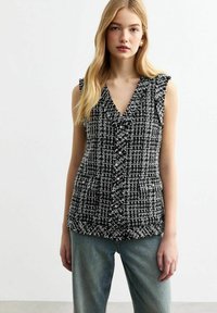 Schwarze und weiße Tweed-Weste mit V-Ausschnitt, gerafften Kanten, Knopfverschluss und zwei vorderen Taschen. Getragen über lockeren blauen Jeans.