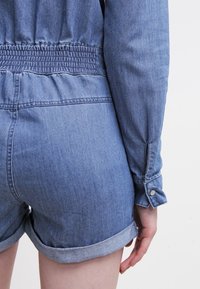 Denim jumpsuit med smockad midja, långa ärmar med knappmanchetter och uppvikt nederkant på shortsen. Blå färg med en mjuk textur.