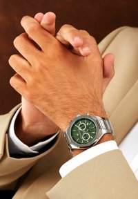 Stalen horloge met een groene textuur wijzerplaat, zilveren accenten en chronograaf functies, gedragen om een gebruinde pols met gevouwen handen.