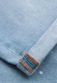 Ljusblå denimtyg med en synlig vävstruktur, med en vikt kant och kontrasterande orange sömnadsdetaljer nära nederkanten.