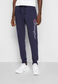 Tmavě modré tepláky z měkké látky s šňůrkou v pase, s bílým vyšitým logem "Champion" na levé straně.