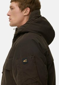 Braune Kapuzenjacke mit einer Fronttasche, strukturiertem Stoff und einem Logopatch am Ärmel. Das Design ist praktisch und utilitaristisch.