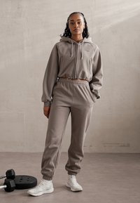 Grijze hoodie trainingspak met een elastische tailleband en manchetten, voorzien van een rits en twee zakken, gecombineerd met witte sportschoenen.