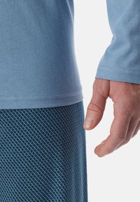Una camisa de manga larga azul clara se superpone a una tela oscura azul con patrón. Primer plano de una mano descansando junto a la cintura de la camisa.
