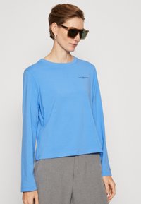 Camicia azzurro chiaro a maniche lunghe in tessuto morbido, con colletto tondo e un piccolo logo nero sul petto, abbinata a pantaloni grigi.