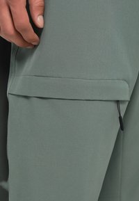 Πράσινο ύφασμα με λεία υφή; διαθέτει μια τσέπη που γ-fold-over με ελαστικό κορδόνι στο στρίφωμα, που τονίζει έναν κομψό, λειτουργικό σχεδιασμό.