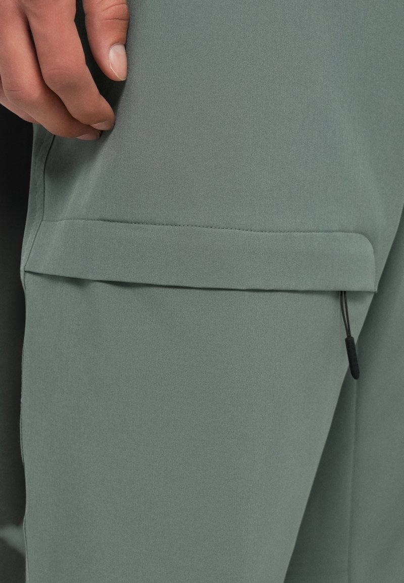 Tissu vert à texture lisse ; présente une poche à rabat avec un cordon élastique à l'ourlet, mettant en avant un design élégant et fonctionnel.