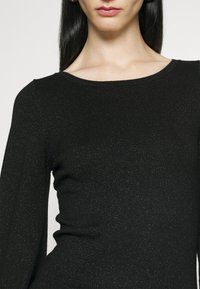 Haut noir à manches longues en tissu texturé à côtes avec un subtil éclat. Présente un décolleté bateau classique et une coupe ajustée.