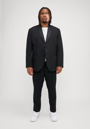 Schwarzer Anzug mit satinierter Spitzlapel, kombiniert mit einem weißen T-Shirt und schwarzen Hosen, ergänzt durch weiße Sneaker. Formelles Design mit Taschen.